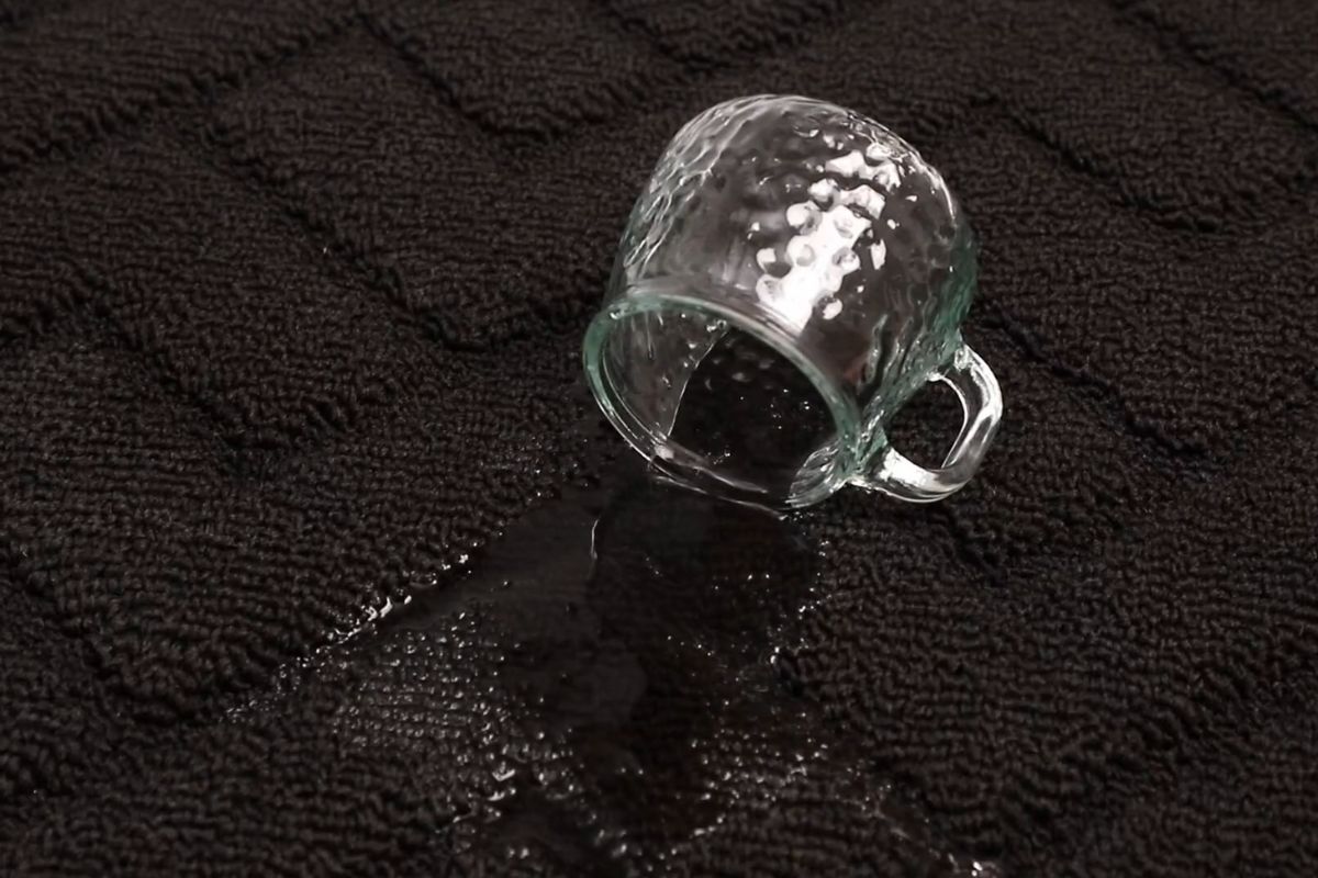 Prevrnuta staklena šolja iz koje se prosula tečnost na tamnom tepihu otpornom na mrlje